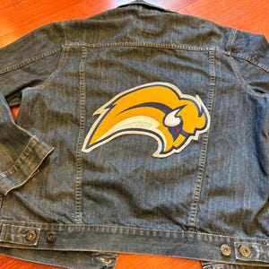 May include: A denim jacket with a large, colorful patch featuring a yellow and blue buffalo logo. The jacket has a classic design with button closures and pockets. The jacket is made of blue denim.