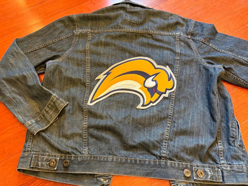 May include: A denim jacket with a large, colorful patch featuring a yellow and blue buffalo logo. The jacket has a classic design with button closures and pockets. The jacket is made of blue denim.