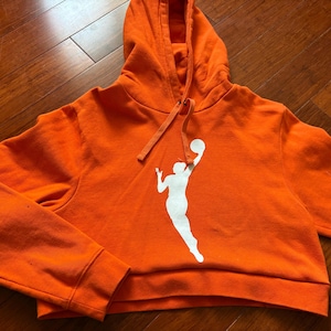 May include: Orange cropped hoodie with a white silhouette of a basketball player dribbling a ball.