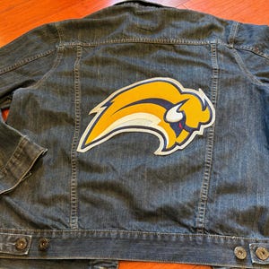 May include: A denim jacket with a large yellow, white, and blue logo patch on the back. The jacket has a classic design with button closures and pockets. The logo is a stylized bison.