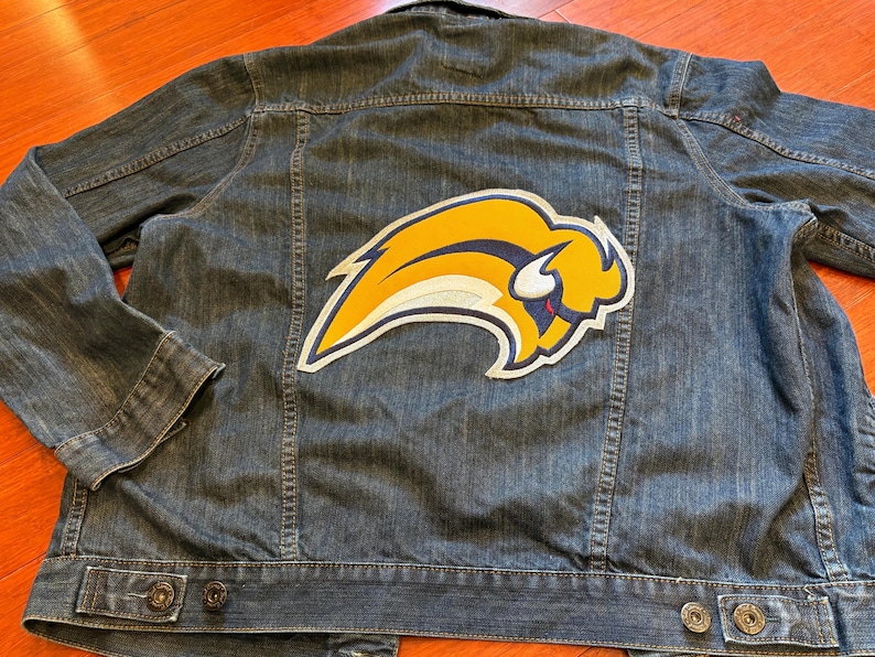 May include: A denim jacket with a large yellow, white, and blue logo patch on the back. The jacket has a classic design with button closures and pockets. The logo is a stylized bison.