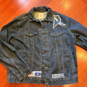May include: A blue denim jacket with a button-up front, two chest pockets, and a collar. The jacket features a logo patch on the upper left chest and a CCM patch on the lower right. Patches with text are visible on the jacket.