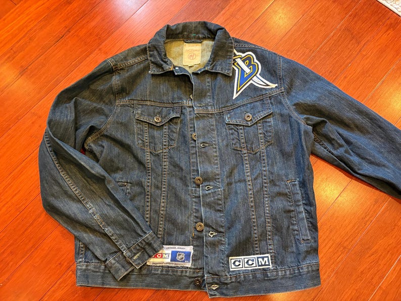 May include: A blue denim jacket with a button-up front, two chest pockets, and a collar. The jacket features a logo patch on the upper left chest and a CCM patch on the lower right. Patches with text are visible on the jacket.