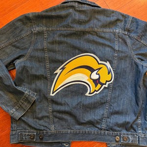 May include: A blue denim jacket with a large yellow and white buffalo logo patch on the back. The jacket has a button-down front, two chest pockets, and two side pockets. The jacket is made of a medium-wash denim material.