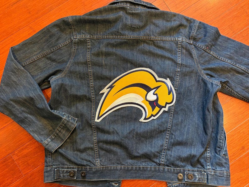 May include: A blue denim jacket with a large yellow and white buffalo logo patch on the back. The jacket has a button-down front, two chest pockets, and two side pockets. The jacket is made of a medium-wash denim material.