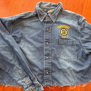 以下が含まれることがあります： 胸にボストン・ブルーインズのロゴが刺繍された、裾がフリンジになった青いデニムシャツです。