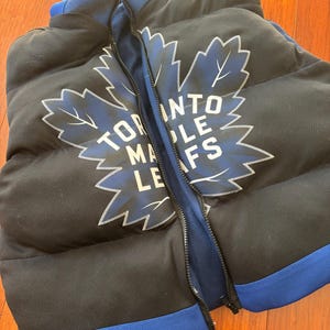 May include: Black and blue puffer vest with a large Toronto Maple Leafs logo on the front. The logo is a white maple leaf with the words "Toronto Maple Leafs" written around it.
