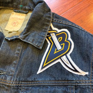 May include: Close-up of a dark blue denim jacket with a large embroidered patch on the upper left chest. The patch features a stylized "B" with a sword, in navy blue, white, and yellow. The jacket has a collar and button closure.