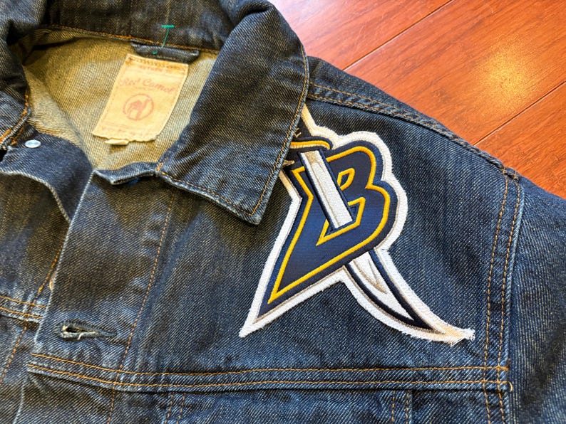 May include: Close-up of a dark blue denim jacket with a large embroidered patch on the upper left chest. The patch features a stylized "B" with a sword, in navy blue, white, and yellow. The jacket has a collar and button closure.