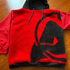 May include: Red and black hooded sweatshirt with a fuzzy texture and a large black graphic on the front.