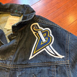 May include: Dark blue denim jacket with a large patch featuring a stylized sword and the letter "B" in navy blue and yellow. The jacket has a classic collar, button closures, and visible stitching.