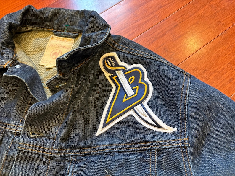 May include: Dark blue denim jacket with a large patch featuring a stylized sword and the letter "B" in navy blue and yellow. The jacket has a classic collar, button closures, and visible stitching.