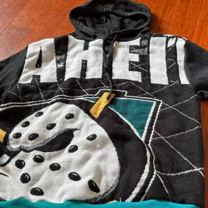 Puede incluir: Sudadera con capucha negra con un diseño temático de hockey. La parte delantera presenta la palabra "MIGHTY" en letras blancas, una máscara de hockey y detalles en turquesa y amarillo. Las mangas son de color negro liso y el dobladillo inferior es turquesa.