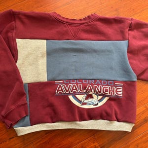May include: A maroon, grey, and blue patchwork sweatshirt with the words "Colorado Avalanche" and a team logo printed on the front.