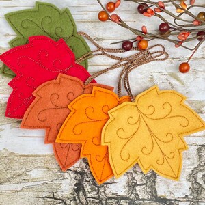 May include: A set of felt leaf ornaments in green, red, orange, and yellow, each featuring decorative brown stitched swirl patterns. The leaves are attached to brown braided cords and are displayed alongside a decorative branch with small berries.