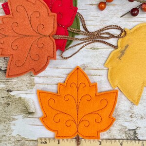 May include: A collection of felt maple leaf ornaments in orange, red, green, and yellow, featuring decorative brown stitched scrollwork patterns. Each leaf is attached to a brown braided cord. A ruler shows the leaves are approximately 10 cm wide.