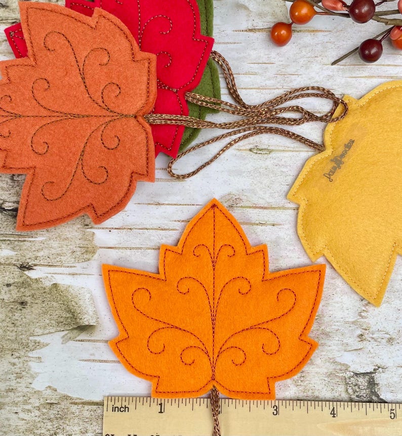 May include: A collection of felt maple leaf ornaments in orange, red, green, and yellow, featuring decorative brown stitched scrollwork patterns. Each leaf is attached to a brown braided cord. A ruler shows the leaves are approximately 10 cm wide.