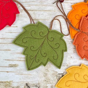 May include: A garland featuring multiple felt maple leaves in shades of red, olive green, orange, and yellow. Each leaf is accented with dark brown decorative swirl stitching and is connected by a thin, metallic copper-coloured cord.
