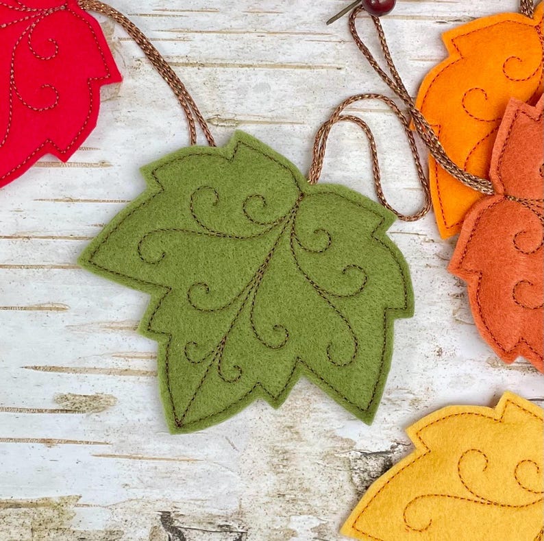 May include: A garland featuring multiple felt maple leaves in shades of red, olive green, orange, and yellow. Each leaf is accented with dark brown decorative swirl stitching and is connected by a thin, metallic copper-coloured cord.