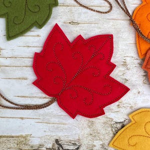 May include: A red felt maple leaf ornament featuring intricate brown stitched swirl patterns and a thin brown cord attached to the base. The leaf is shaped with multiple points and is part of a collection of similar felt leaf decorations.