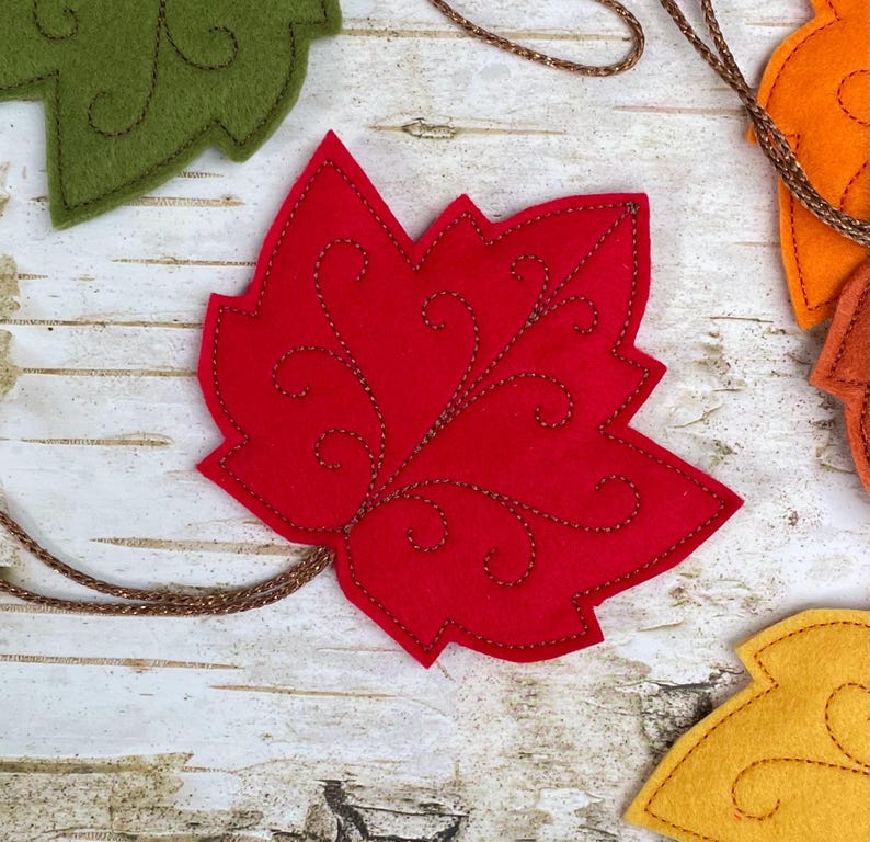 May include: A red felt maple leaf ornament featuring intricate brown stitched swirl patterns and a thin brown cord attached to the base. The leaf is shaped with multiple points and is part of a collection of similar felt leaf decorations.