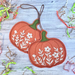 May include: Two orange felt pumpkin ornaments with green stems and white floral embroidery. Each pumpkin has a brown string for hanging. The pumpkins are on a light wood surface with colorful paper shreds.