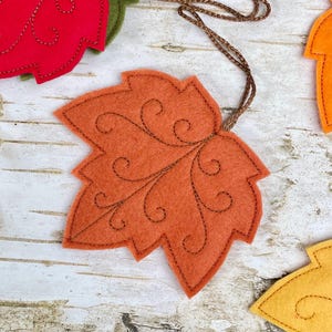 May include: A felt maple leaf ornament in a vibrant orange hue, featuring intricate dark brown stitched swirl patterns across its surface. The leaf is attached to a thin, braided brown cord for hanging, showcasing detailed craftsmanship.