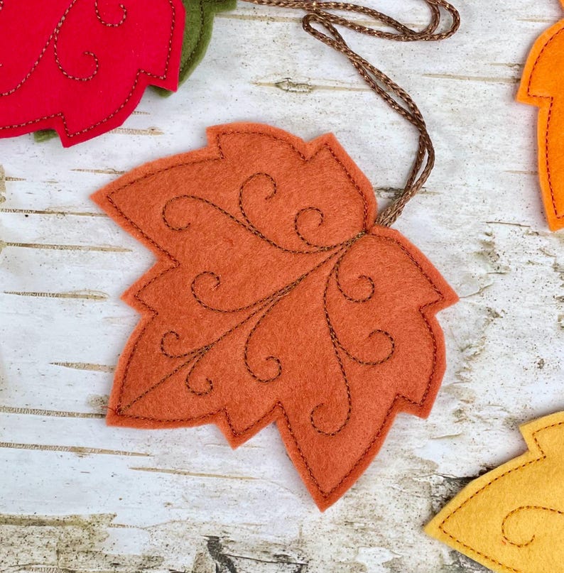 May include: A felt maple leaf ornament in a vibrant orange hue, featuring intricate dark brown stitched swirl patterns across its surface. The leaf is attached to a thin, braided brown cord for hanging, showcasing detailed craftsmanship.