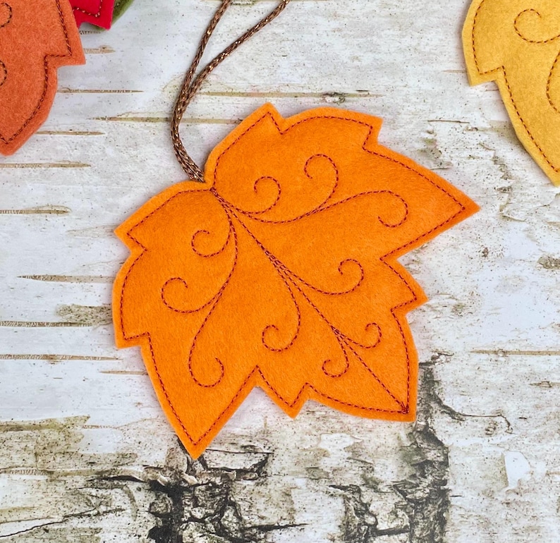 May include: An orange felt maple leaf ornament featuring dark orange stitched swirl patterns and a brown metallic hanging cord. The leaf has a jagged edge design and is crafted from soft fabric material, designed to be hung as a decorative item.
