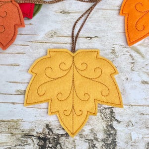 May include: A mustard yellow felt leaf ornament featuring dark brown stitched scrollwork details and a central vein. The leaf is attached to a thin, metallic brown hanging cord, with additional orange and red felt leaves visible nearby.
