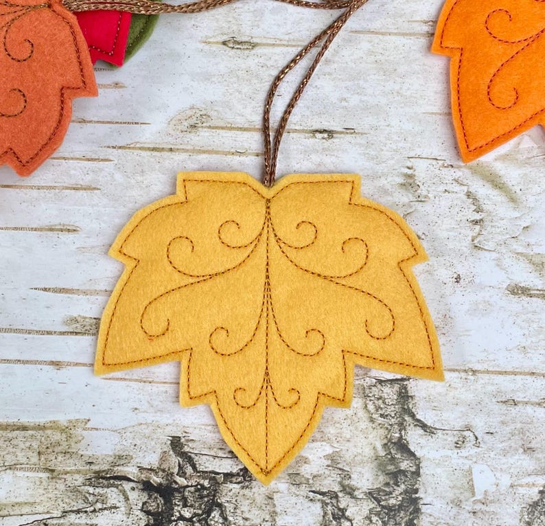 May include: A mustard yellow felt leaf ornament featuring dark brown stitched scrollwork details and a central vein. The leaf is attached to a thin, metallic brown hanging cord, with additional orange and red felt leaves visible nearby.