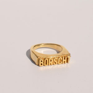 Peut inclure: Une bague dorée avec le mot "BORSCHT" en majuscules. La bague a une face rectangulaire et une finition lisse et polie. La bague est posée sur un fond blanc.