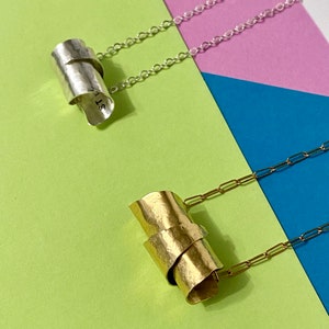 May include: Two silver and gold necklaces with unique, abstract pendant designs. The silver pendant is a twisted, hammered metal tube. The gold pendant is a twisted, hammered metal tube. Both pendants are hanging from delicate chain necklaces.