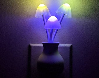 May include: A white ceramic night light shaped like a vase with three mushroom-shaped lights. The lights are glowing in different colors: yellow, purple, and blue.