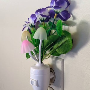 May include: A white night light shaped like a flower pot with purple and white flowers and mushroom-shaped lights. The night light is plugged into a standard electrical outlet.