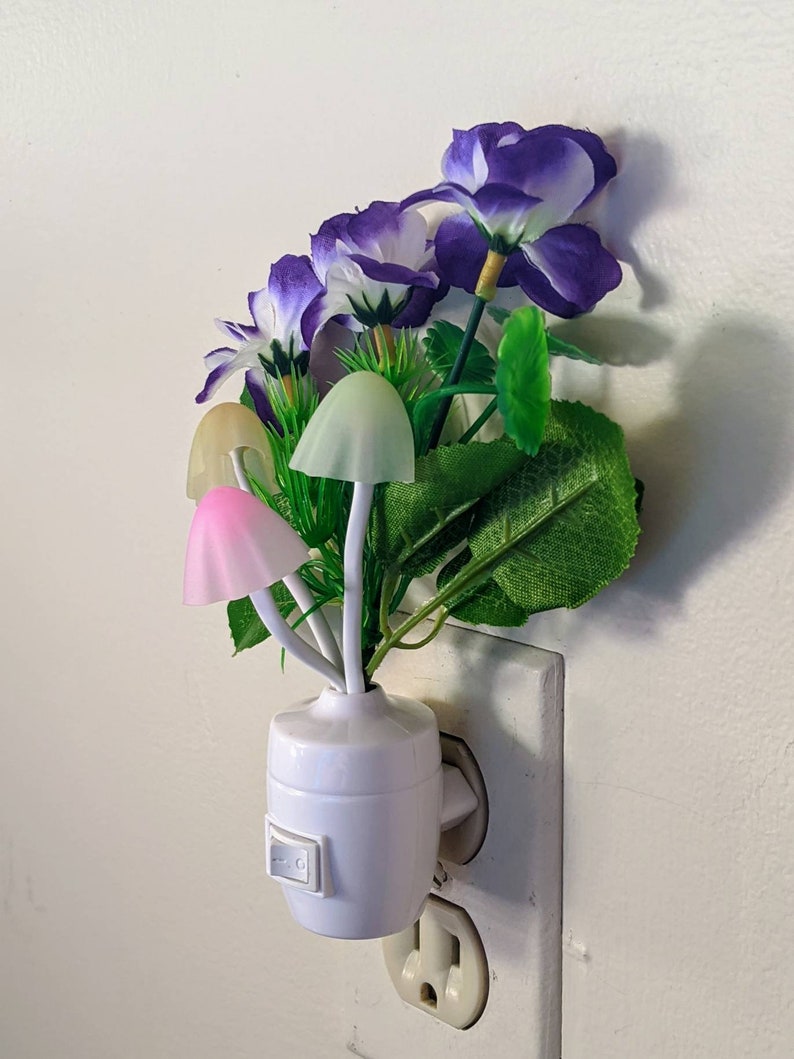 May include: A white night light shaped like a flower pot with purple and white flowers and mushroom-shaped lights. The night light is plugged into a standard electrical outlet.