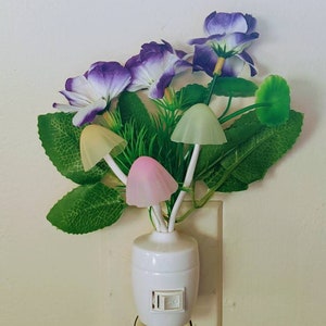 May include: A white night light shaped like a mushroom with a purple flower arrangement. The night light is plugged into an electrical outlet.