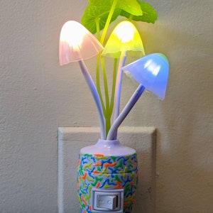 May include: A white mushroom-shaped night light with a colorful base and a switch. The night light has three mushroom heads that glow in different colors: pink, yellow, and blue. The base is decorated with a colorful swirl pattern.