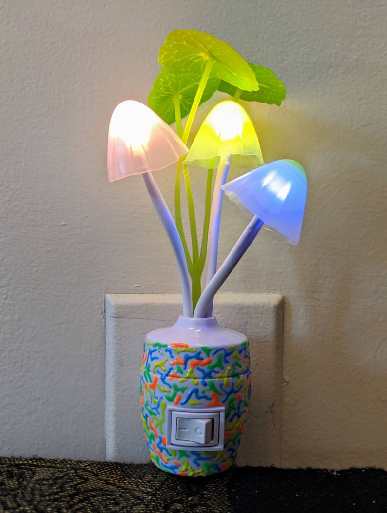 May include: A white mushroom-shaped night light with a colorful base and a switch. The night light has three mushroom heads that glow in different colors: pink, yellow, and blue. The base is decorated with a colorful swirl pattern.