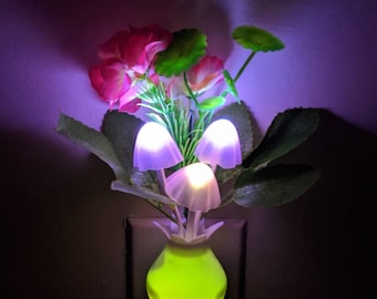 May include: A green and white night light shaped like a vase with three purple mushroom-shaped lights. The vase is plugged into an electrical outlet and has pink flowers coming out of the top.