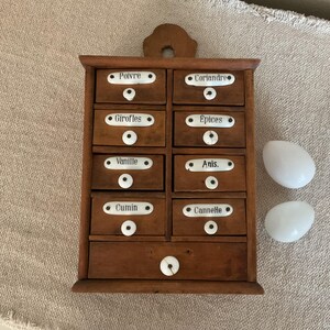 May include: A wooden spice cabinet with eight small drawers and one larger bottom drawer, each featuring white ceramic labels with black text including Poivre, Coriandre, Girofles, Epices, Vanille, Anis, Cumin, and Cannelle, plus two white oval stones.