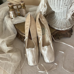 May include: A pair of vintage satin ballet slippers in a cream color, with delicate ribbon ties and small bows. The shoes are displayed on a light-colored surface, with a sheer fabric and decorative items in the background.