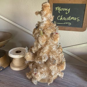 May include: A tabletop Christmas tree made of light beige tinsel with a textured, fuzzy appearance. The tree is decorated with several small, clear, and colored plastic light bulbs inserted into the branches throughout the structure.