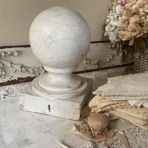 May include: A distressed white sphere finial on a square base, with a keyhole detail. The finial sits on a table with vintage lace and a small hat with flowers. A basket of dried flowers is in the background.