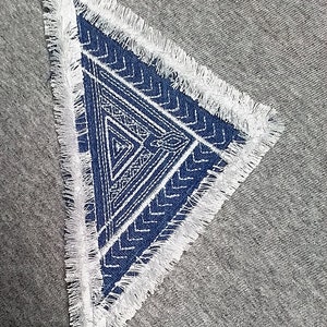 May include: A blue triangle patch with a white geometric pattern and a white frayed border. The patch is sewn onto a gray fabric background.
