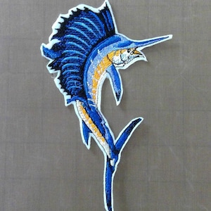 May include: Embroidered iron-on patch of a blue marlin with yellow and white accents. The marlin is jumping out of the water with its dorsal fin extended.