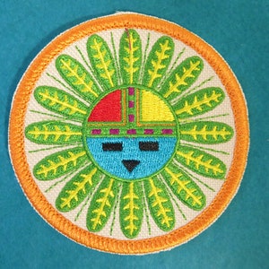 May include: A round embroidered patch with an orange border. The center of the patch features a stylized face with a blue background, red, yellow, and purple accents, and green and yellow leaves radiating outward.