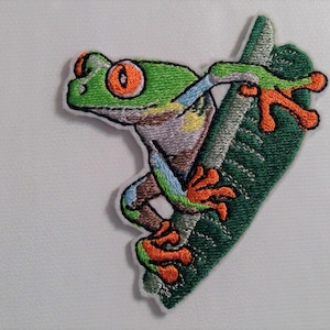 May include: Embroidered iron-on patch of a green tree frog with orange feet, climbing a green leaf.