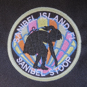 May include: Embroidered patch with the words "SANIBEL ISLAND" and "SANIBEL STOOP" in white lettering. The patch features a black silhouette of a person stooping, with a colorful, segmented background in shades of pink, orange, yellow, and blue.