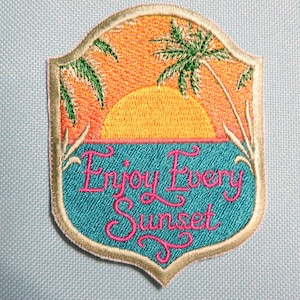 May include: Embroidered patch with a sunset scene. The design features a yellow sun, orange sky, blue water, and green palm trees. The text "Enjoy Every Sunset" is in pink script. The patch has a light blue background.